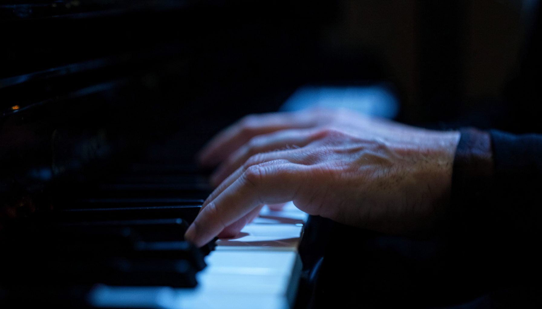 Primer plano de las manos de un pianista sobre teclas iluminadas por una luz azul tenue, con el resto del escenario en penumbra