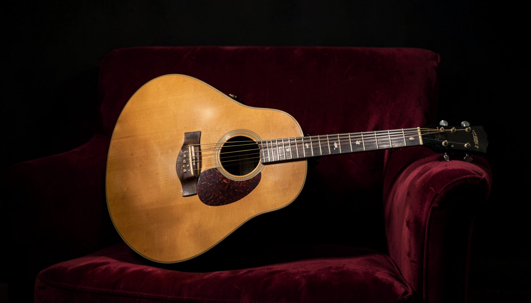 Una guitarra acústica apoyada sobre un sillón de terciopelo granate, iluminada por una luz cálida y tenue.