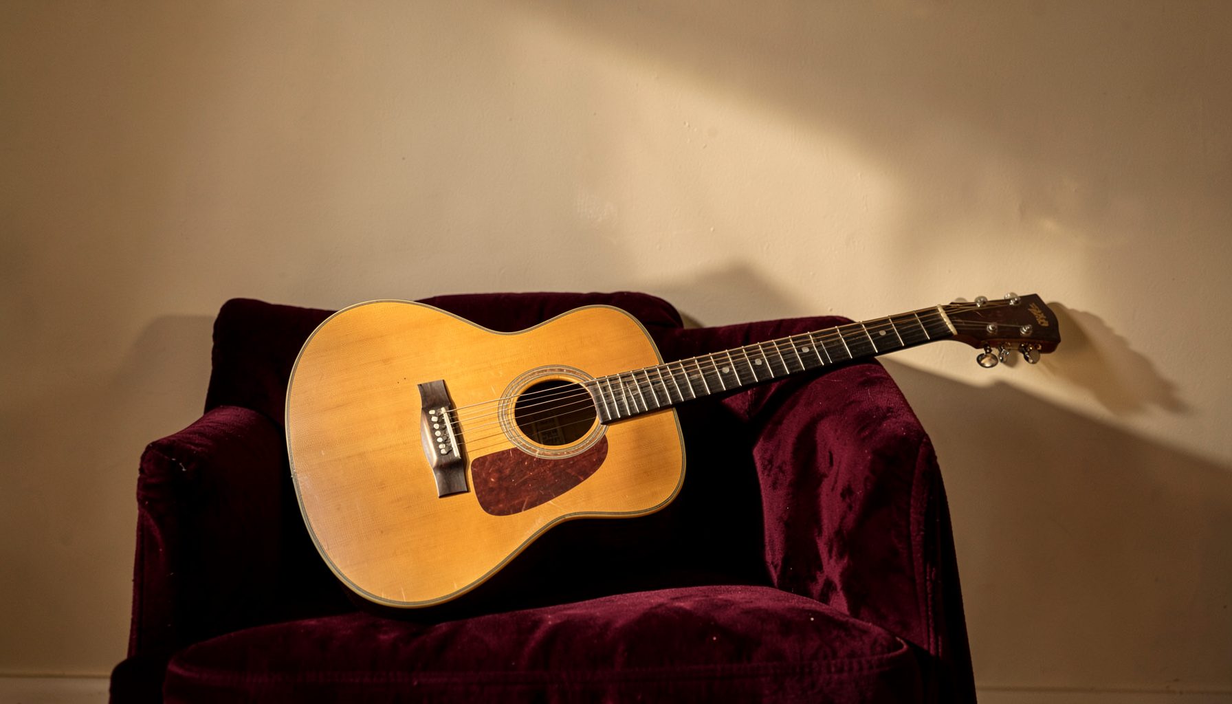 Una guitarra acústica apoyada sobre un sillón de terciopelo granate, iluminada por una luz cálida y tenue que proyecta sombras suaves.